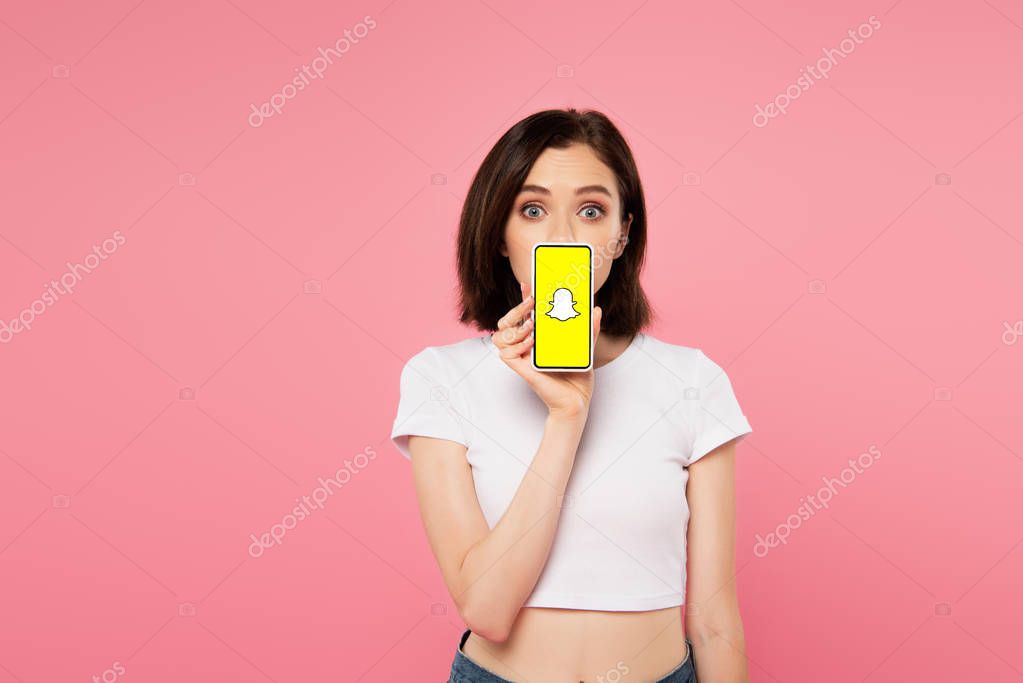 KYIV, UKRAINE - JULY 3, 2019: shocked girl holding smartphone with snapchat logo isolated on pink