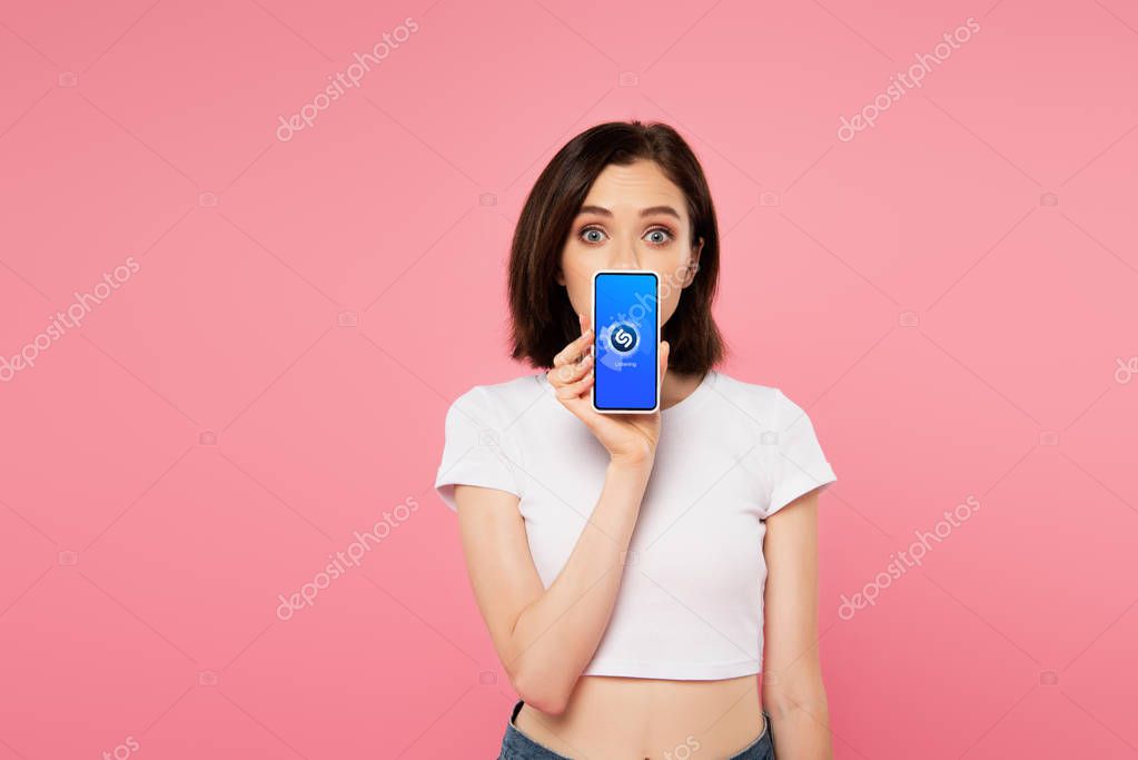 KYIV, UKRAINE - JULY 3, 2019: shocked girl holding smartphone with shazam logo isolated on pink