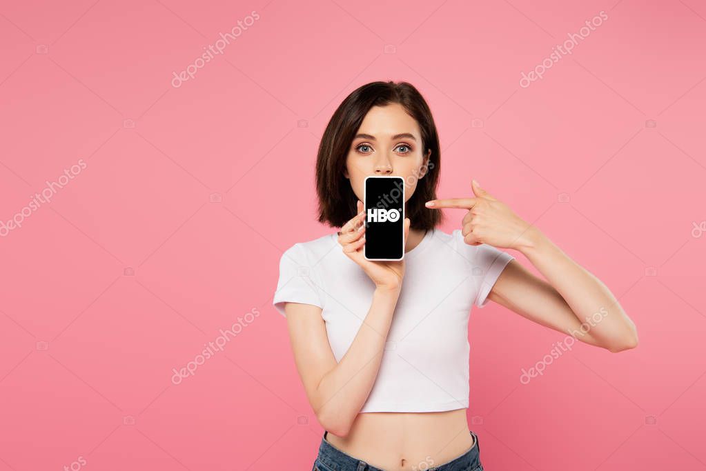 KYIV, UKRAINE - JULY 3, 2019: shocked girl pointing with finger at smartphone with HBO logo isolated on pink