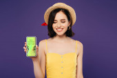 usměvavá dívka v slaměném klobouku držící telefon Smartphone s beat Shopping App izolovaný na fialový