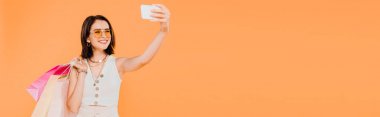 alışveriş çantaları ile güneş gözlüğü mutlu moda kız turuncu izole akıllı telefonda selfie alarak