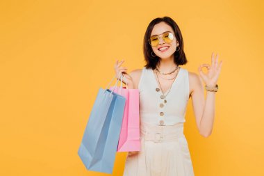 happy fashionable girl in sunglasses holding shopping bags and showing okay sign isolated on yellow clipart