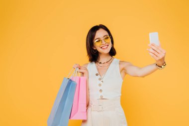 alışveriş çantaları ile güneş gözlüğü mutlu moda kız sarı izole akıllı telefonda selfie alarak