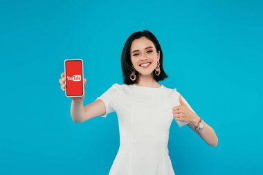 Kiev, Ukrayna - 3 Temmuz 2019: youtube logolu akıllı telefonu tutan ve mavi üzerinde izole başparmak gösteren elbise zarif kadın gülümseyen
