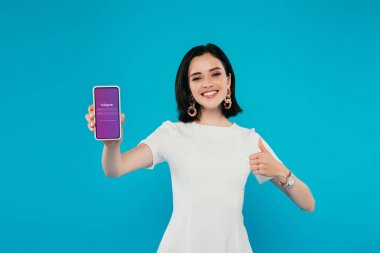 Kiev, Ukrayna - 3 Temmuz 2019: Instagram logolu akıllı telefonu tutan ve mavi üzerinde izole başparmak gösteren elbise zarif kadın gülümseyen