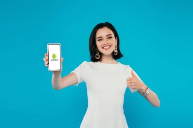 Kiev, Ukrayna - 3 Temmuz 2019: android logolu akıllı telefonu tutan ve mavi üzerinde izole başparmak gösteren elbise zarif kadın gülümseyen