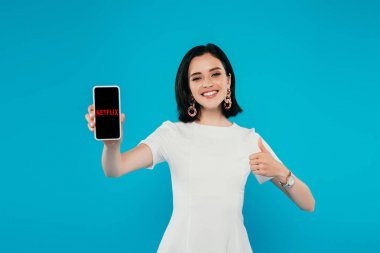 Kiev, Ukrayna - 3 Temmuz 2019: Netflix logolu akıllı telefonu tutan ve mavi üzerinde izole başparmak gösteren elbise zarif kadın gülümseyen