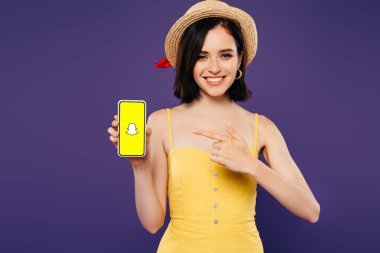 KYIV, UKRAINE - JULY 3, 2019: smiling girl in straw hat pointing with finger at smartphone with snapchat app isolated on purple clipart
