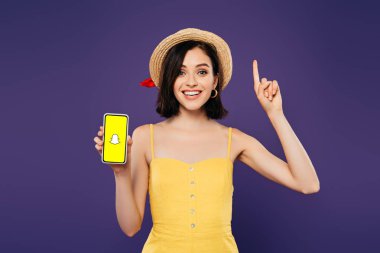 Kiev, Ukrayna - 3 Temmuz 2019: hasır şapkalı heyecanlı kız fikir jesti gösteriyor ve mor üzerinde izole snapchat uygulaması ile akıllı telefon tutuyor