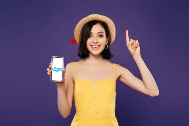 Kiev, Ukrayna - 3 Temmuz 2019: hasır şapkalı heyecanlı kız fikir jesti gösteriyor ve mor üzerinde izole skype uygulaması ile akıllı telefon tutuyor