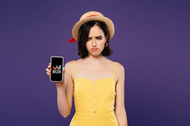 Kiev, Ukrayna - 3 Temmuz 2019: hasır şapkalı üzgün güzel kız mor üzerinde izole deezer uygulaması ile akıllı telefon tutan 