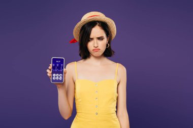 hasır şapka lı üzgün güzel kız, mor üzerinde izole sağlık uygulaması ile akıllı telefon tutuyor 