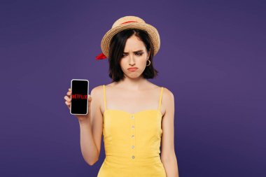 KYIV, UKRAINE - JULY 3, 2019: sad pretty girl in straw hat holding smartphone with netflix app isolated on purple  clipart