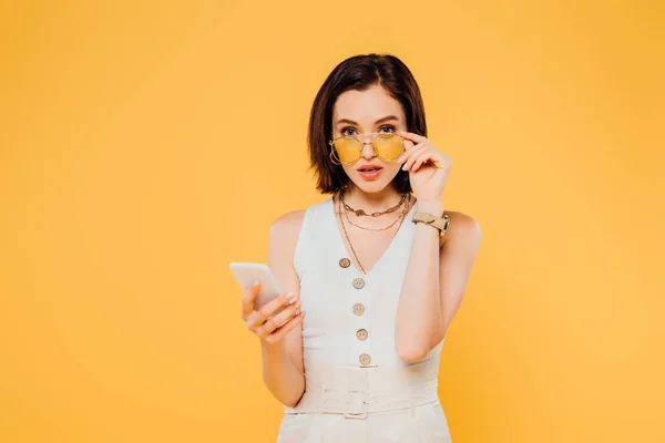 Wstrząśnięty Kobieta Okulary Przeciwsłoneczne Trzymając Smartfon Dotykając Okulary Izolowane Żółto — Zdjęcie stockowe
