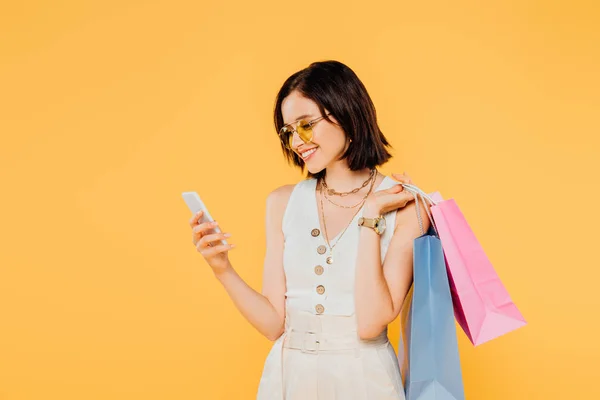 微笑时尚女孩在太阳镜与购物袋使用智能手机隔离在黄色 — 图库照片