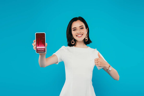 smiling elegant woman in dress holding smartphone with trading courses app and showing thumb up isolated on blue