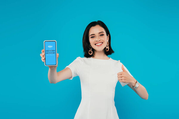 KYIV, UKRAINE - JULY 3, 2019: smiling elegant woman in dress holding smartphone with skype logo and showing thumb up isolated on blue
