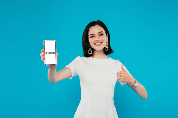 KYIV, UKRAINE - JULY 3, 2019: smiling elegant woman in dress holding smartphone with tinder logo and showing thumb up isolated on blue