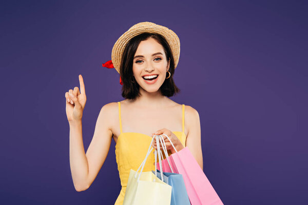 excited smiling girl in straw hat with shopping bags showing idea sign isolated on purple