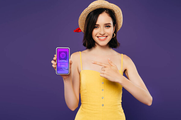smiling girl in straw hat pointing with finger at smartphone with online shopping app isolated on purple