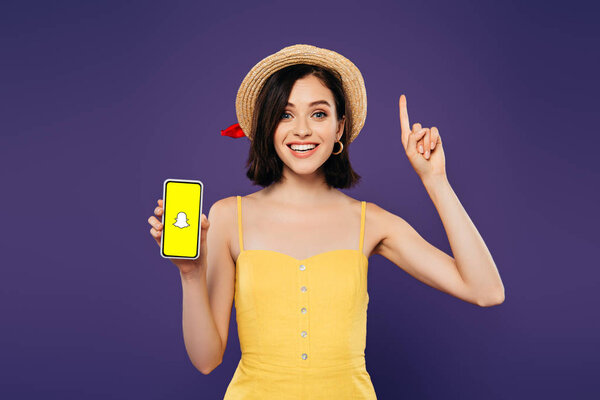 KYIV, UKRAINE - JULY 3, 2019: excited girl in straw hat showing idea gesture and holding smartphone with snapchat app isolated on purple