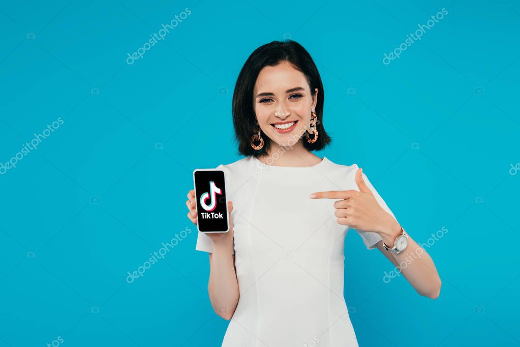 KYIV, UKRAINE - JULY 3, 2019: smiling elegant woman in dress pointing with finger at smartphone with tiktok logo isolated on blue