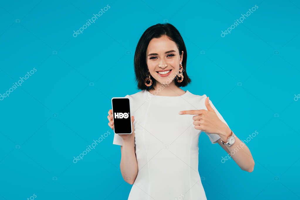 KYIV, UKRAINE - JULY 3, 2019: smiling elegant woman in dress pointing with finger at smartphone with HBO logo isolated on blue