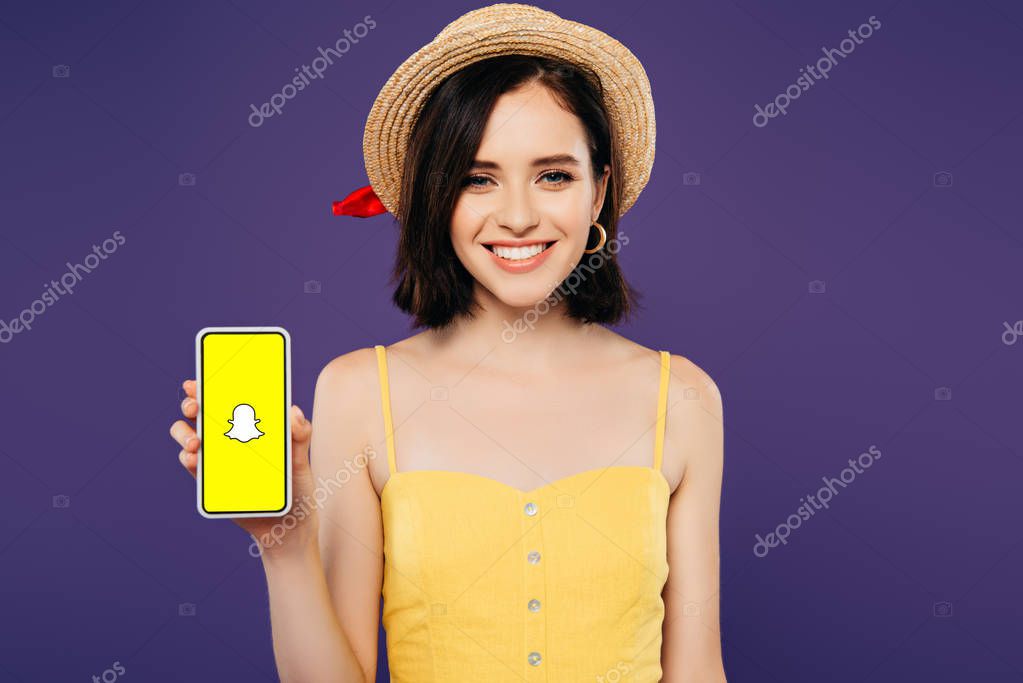 KYIV, UKRAINE - JULY 3, 2019: smiling girl in straw hat holding smartphone with Snapchat app isolated on purple