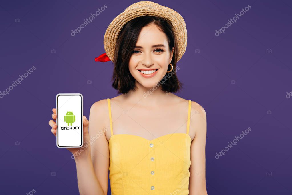 KYIV, UKRAINE - JULY 3, 2019: smiling girl in straw hat holding smartphone with android logo isolated on purple