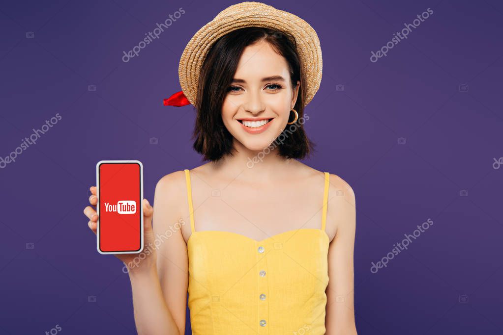 KYIV, UKRAINE - JULY 3, 2019: smiling girl in straw hat holding smartphone with youtube app isolated on purple