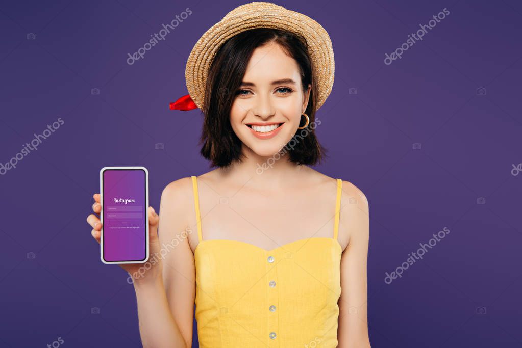 KYIV, UKRAINE - JULY 3, 2019: smiling girl in straw hat holding smartphone with Instagram app isolated on purple