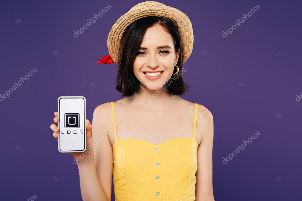 KYIV, UKRAINE - JULY 3, 2019: smiling girl in straw hat holding smartphone with uber app isolated on purple