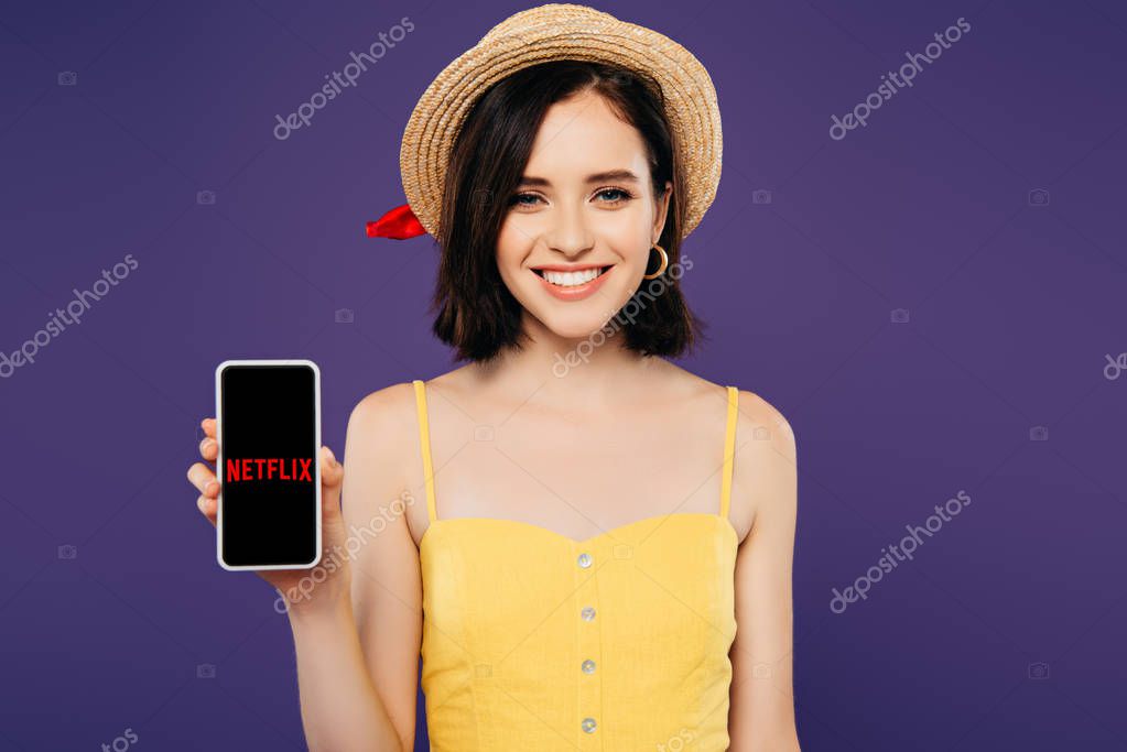 KYIV, UKRAINE - JULY 3, 2019: smiling girl in straw hat holding smartphone with netflix app isolated on purple