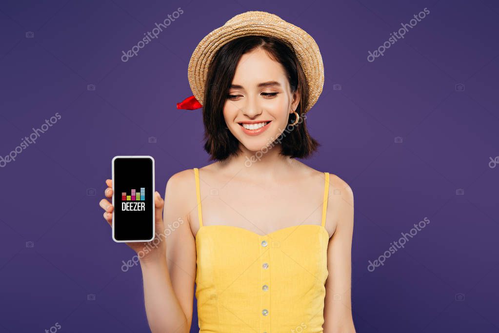 KYIV, UKRAINE - JULY 3, 2019: smiling girl in straw hat holding smartphone with deezer app isolated on purple