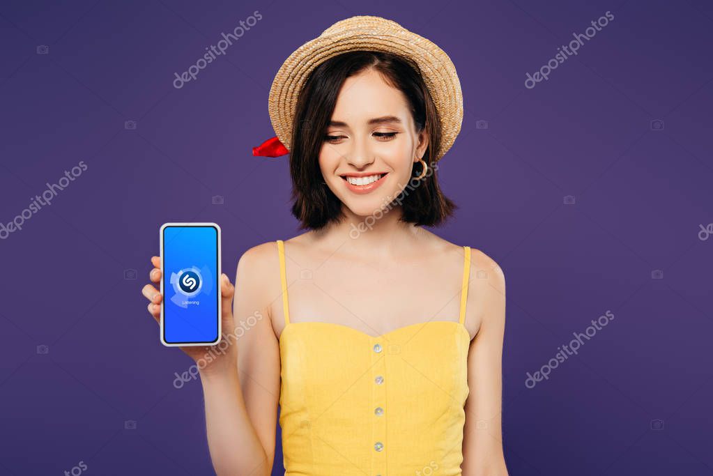 KYIV, UKRAINE - JULY 3, 2019: smiling girl in straw hat holding smartphone with shazam app isolated on purple