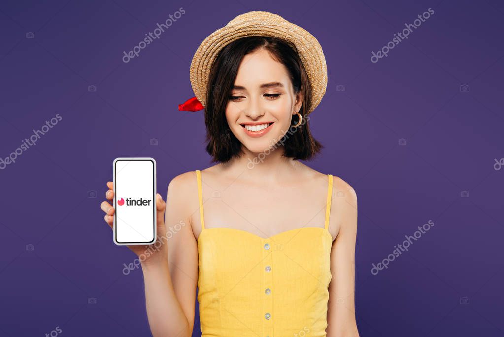 KYIV, UKRAINE - JULY 3, 2019: smiling girl in straw hat holding smartphone with tinder app isolated on purple