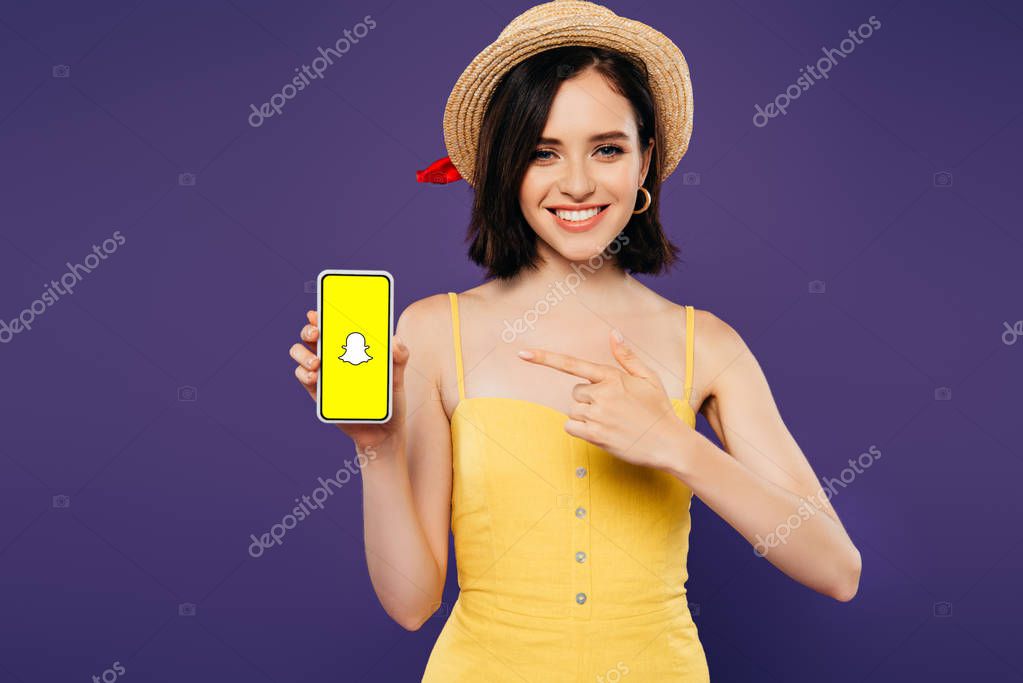 KYIV, UKRAINE - JULY 3, 2019: smiling girl in straw hat pointing with finger at smartphone with snapchat app isolated on purple