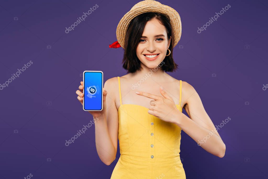KYIV, UKRAINE - JULY 3, 2019: smiling girl in straw hat pointing with finger at smartphone with shazam app isolated on purple