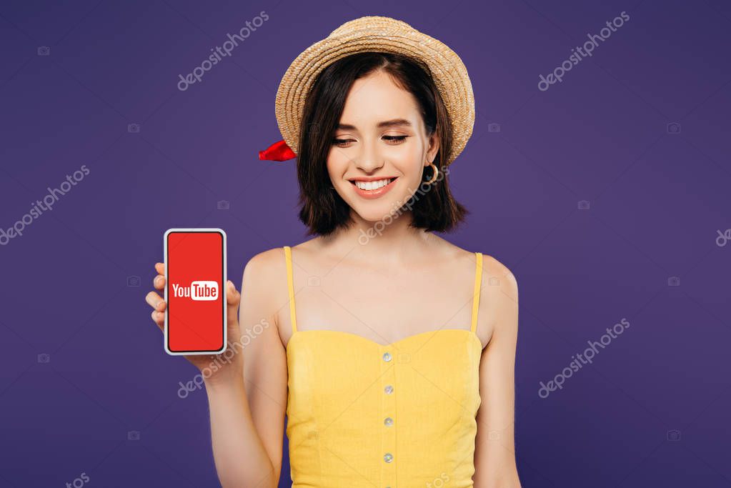 KYIV, UKRAINE - JULY 3, 2019: smiling girl in straw hat holding smartphone with youtube app isolated on purple
