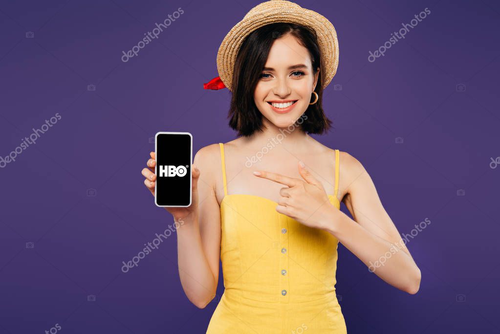 KYIV, UKRAINE - JULY 3, 2019: smiling girl in straw hat pointing with finger at smartphone with HBO app isolated on purple