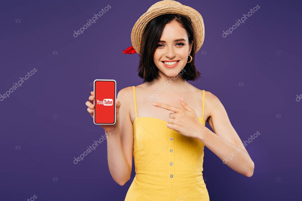 KYIV, UKRAINE - JULY 3, 2019: smiling girl in straw hat pointing with finger at smartphone with youtube app isolated on purple