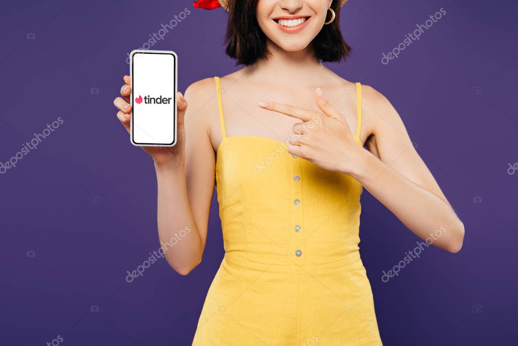 KYIV, UKRAINE - JULY 3, 2019: cropped view of girl in straw hat pointing with finger at smartphone with tinder app isolated on purple