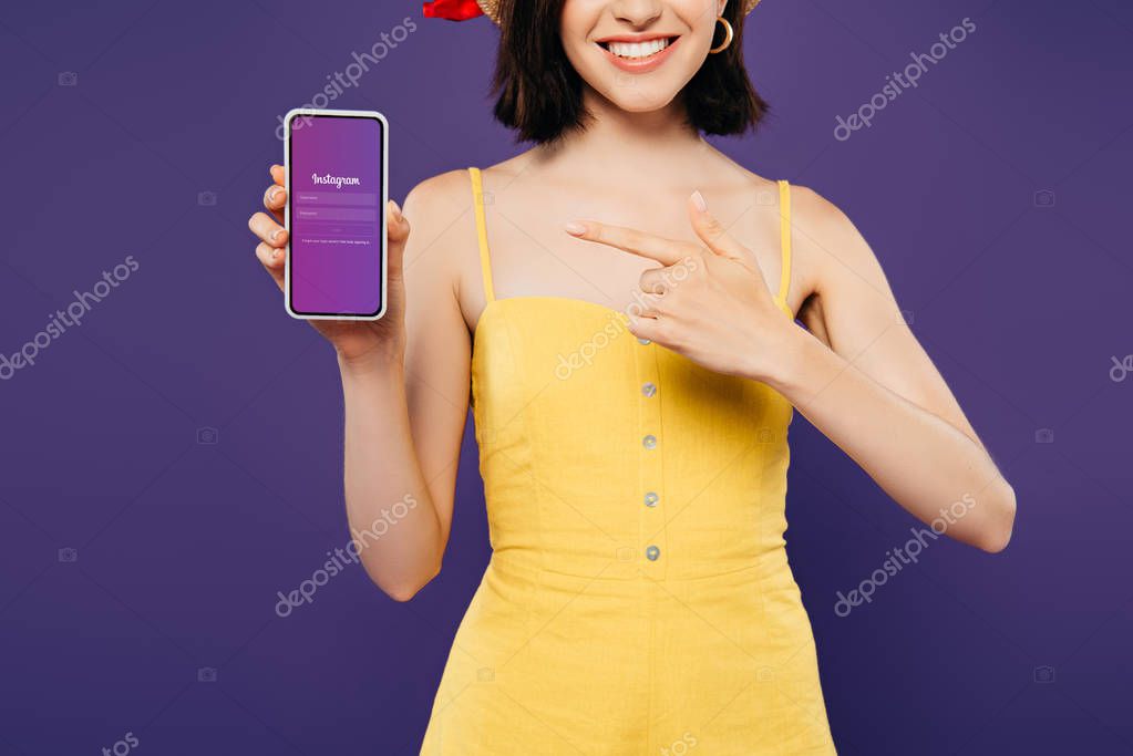 KYIV, UKRAINE - JULY 3, 2019: cropped view of girl in straw hat pointing with finger at smartphone with instagram app isolated on purple
