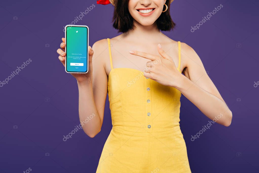 KYIV, UKRAINE - JULY 3, 2019: cropped view of girl in straw hat pointing with finger at smartphone with twitter app isolated on purple
