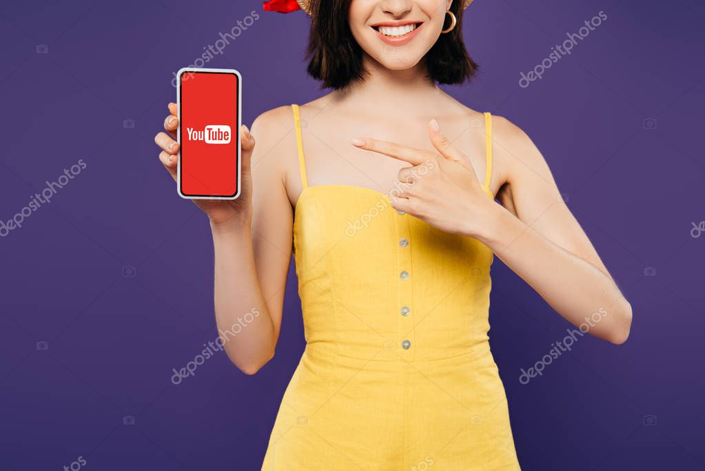 KYIV, UKRAINE - JULY 3, 2019: cropped view of girl in straw hat pointing with finger at smartphone with youtube app isolated on purple