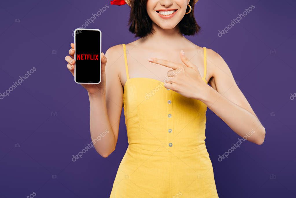 KYIV, UKRAINE - JULY 3, 2019: cropped view of girl in straw hat pointing with finger at smartphone with netflix app isolated on purple