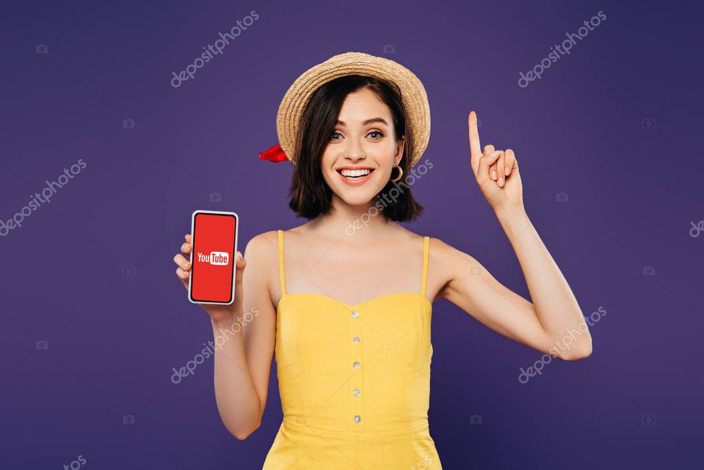 KYIV, UKRAINE - JULY 3, 2019: excited girl in straw hat showing idea gesture and holding smartphone with youtube app isolated on purple