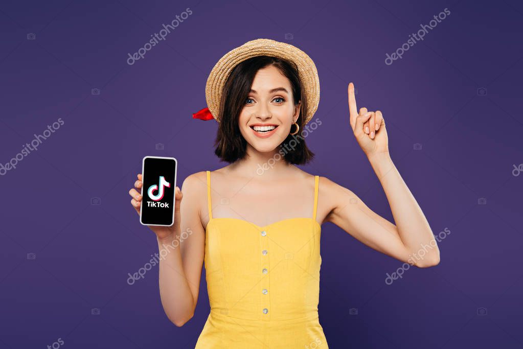 KYIV, UKRAINE - JULY 3, 2019: excited girl in straw hat showing idea gesture and holding smartphone with tiktok app isolated on purple