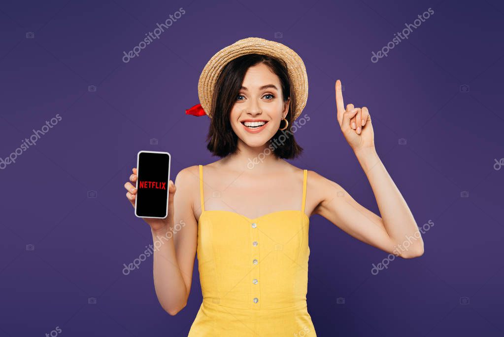 KYIV, UKRAINE - JULY 3, 2019: excited girl in straw hat showing idea gesture and holding smartphone with netflix app isolated on purple