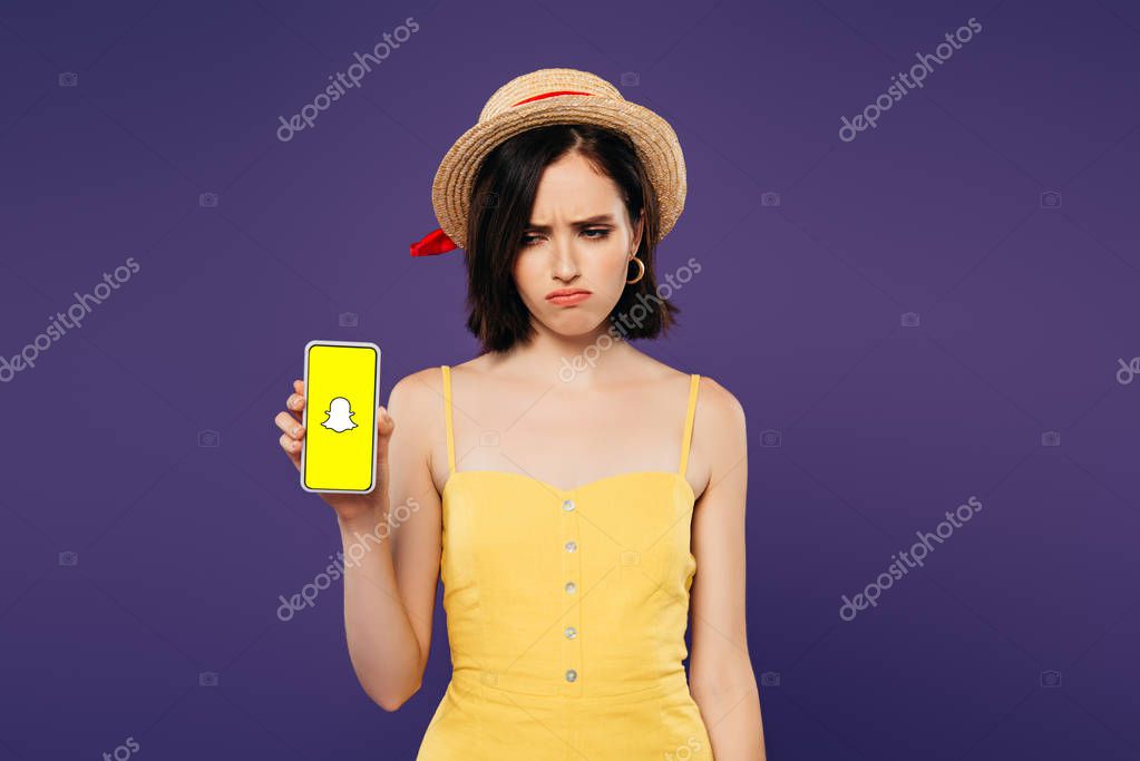 KYIV, UKRAINE - JULY 3, 2019: sad pretty girl in straw hat holding smartphone with snapchat app isolated on purple
